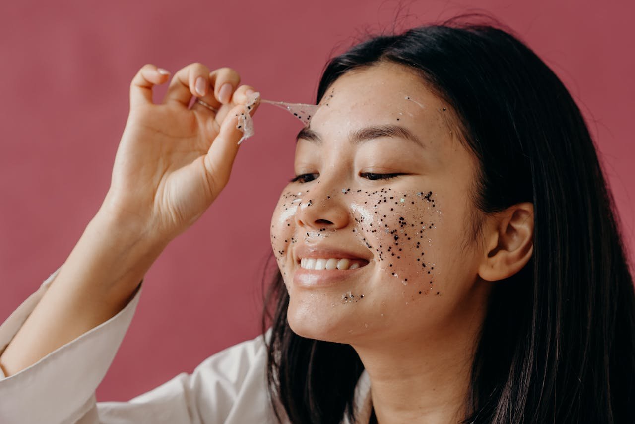 hero-gallery-01 Close-up of smiling Asian woman peeling off a glittery face mask, highlighting her skincare routine.