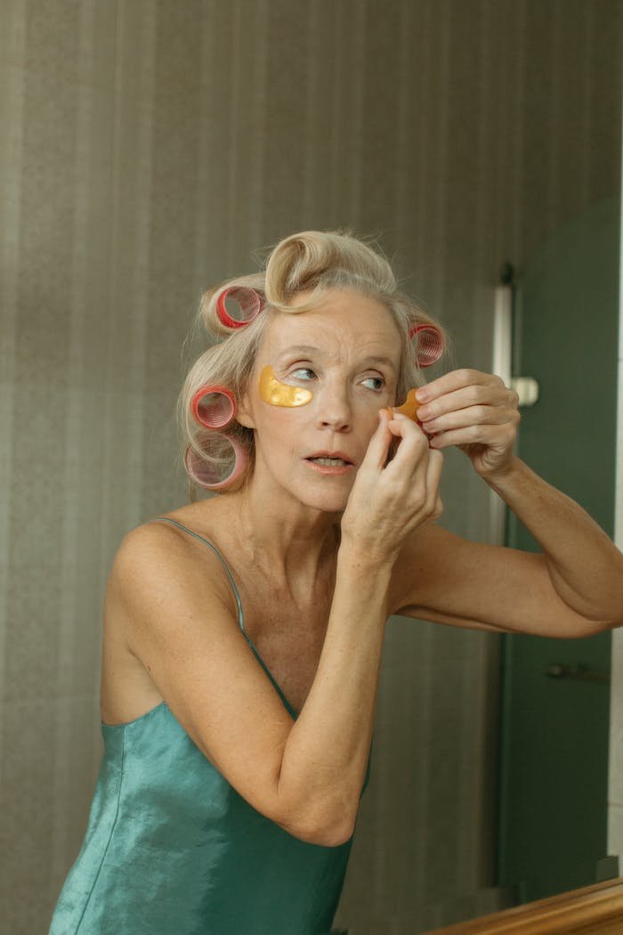 hero-gallery-03 Elderly woman applies skincare patches with hair in curlers indoors.